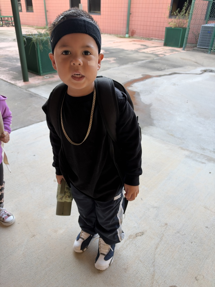 Child wearing a black jacket, pants, and shoes, with a backpack and gold necklace, stands on concrete.