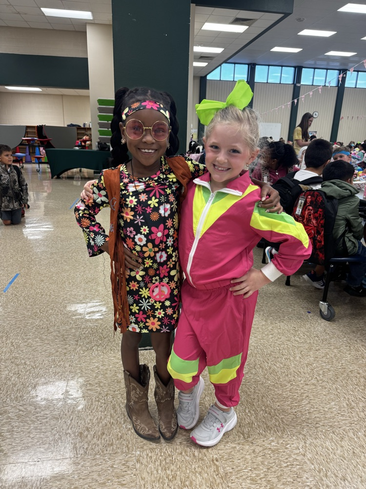 Two young girls in costumes, one with a floral dress and the other in a pink jumpsuit.