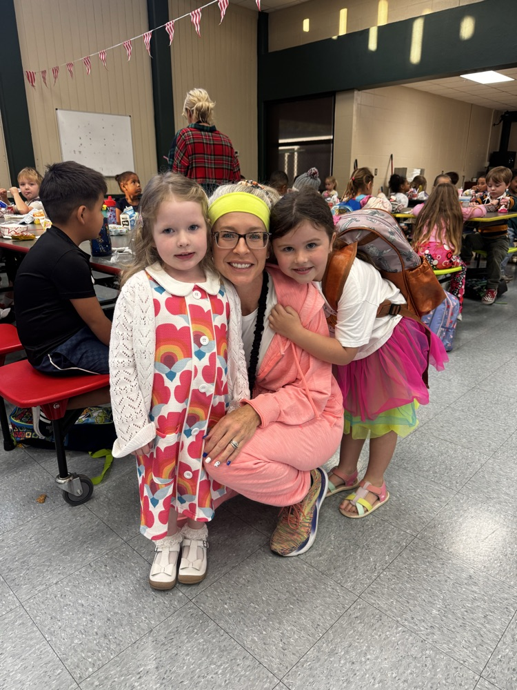 Woman and two young girls posing in a school cafeteria with tables and chairs in background.