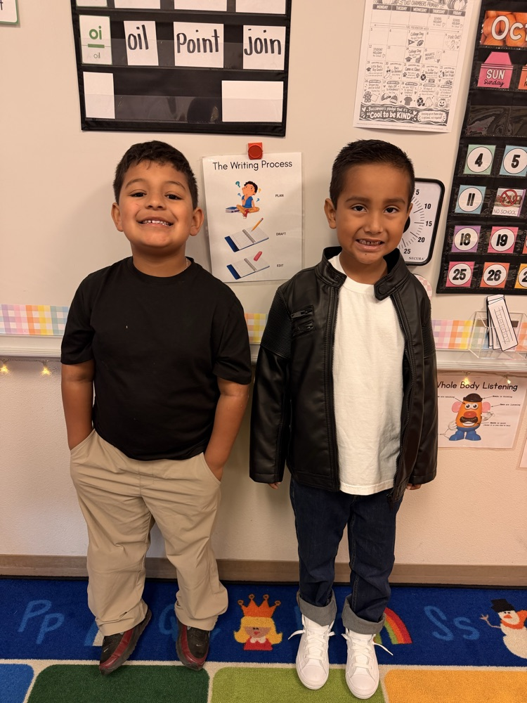 Two young boys smiling, standing side by side on a colorful classroom rug with letters on the floor.