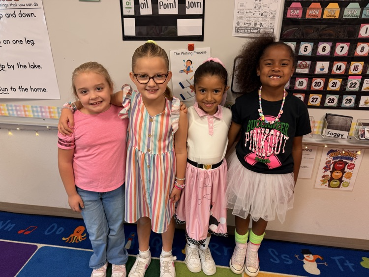 Four girls stand together, smiling, with one girl wearing glasses and another wearing a necklace.
