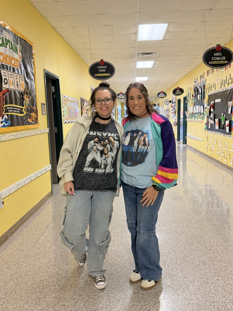 Two people stand in a yellow hallway, smiling and wearing glasses. The hallway has posters and a ceiling with lights.