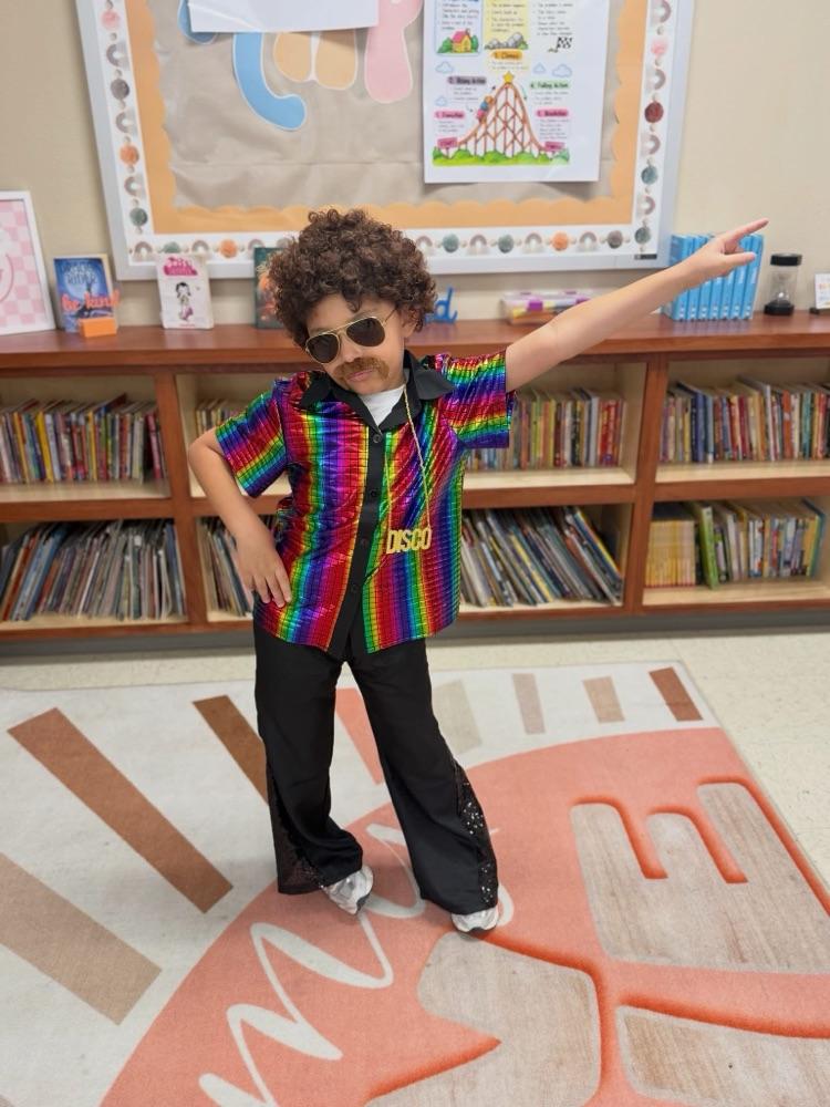 A person in a colorful disco outfit and sunglasses stands with arm outstretched in front of bookshelf.