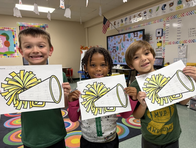 Three children hold up drawings of yellow pom-poms and megaphones in a classroom.