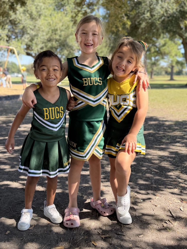 Three girls in cheerleading uniforms smile for the camera in a park with playground in background.