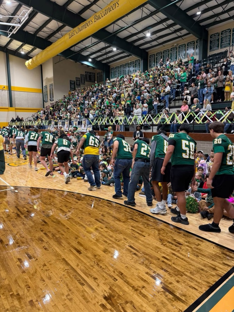 Several adults in green jerseys line up on a basketball court; a crowd of people sits in the bleachers.