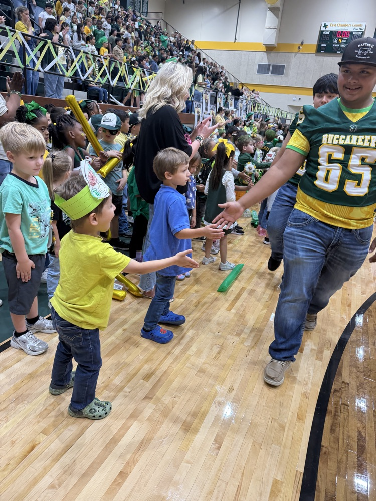 Several children wearing hats and sneakers in a gym, one child wearing a green hat shakes hands with a man in a football jersey.