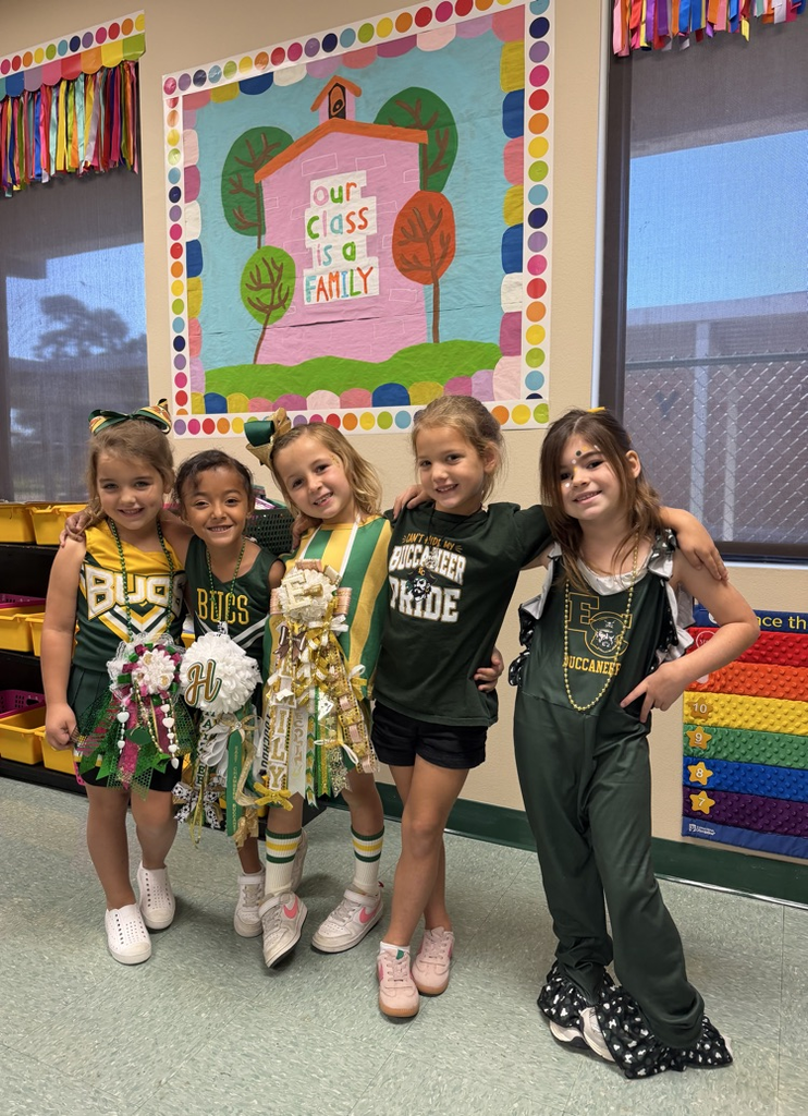 Five girls in cheerleading uniforms standing together in front of a classroom display.