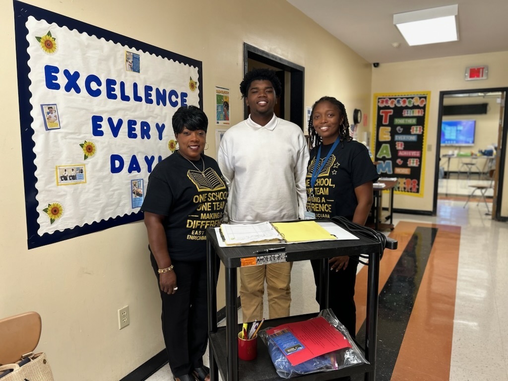 A board member, a student, and a teacher in front of a cart at EFEA and a banner that says excellence every day.