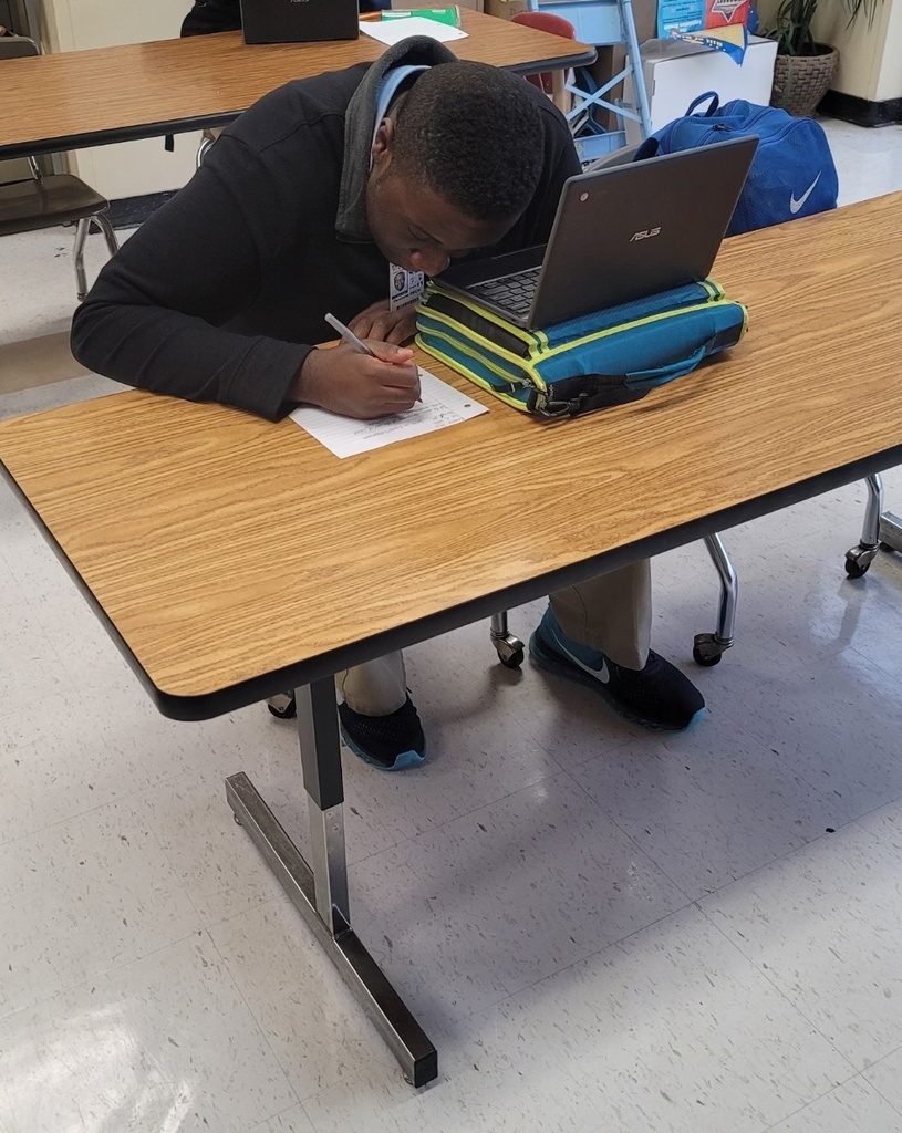 A student is writing by hand next to an open Chromebook.