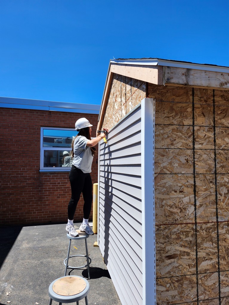 student hammers siding on shed with construction hat and glasses on