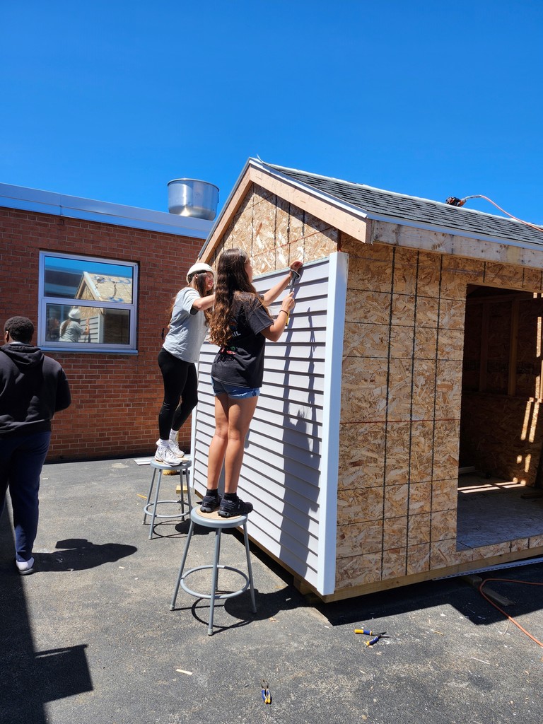 Students hammer gray siding on their shed