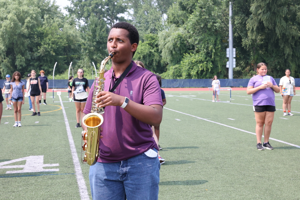 Student plays the saxophone with a purple shirt and jeans on the turf football field
