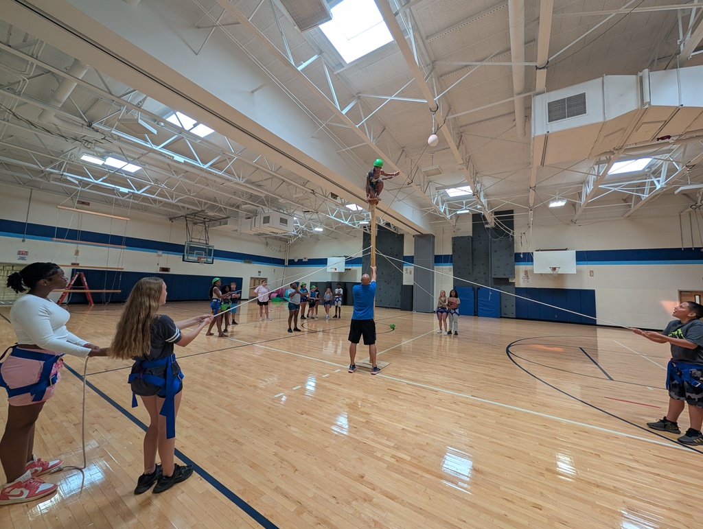 Student stands on pole on adventure course wide shot and other studens are holding rope to support the structure