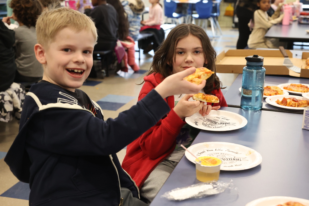 two students in a busy cafeteria hold up pizza and smile for a photo