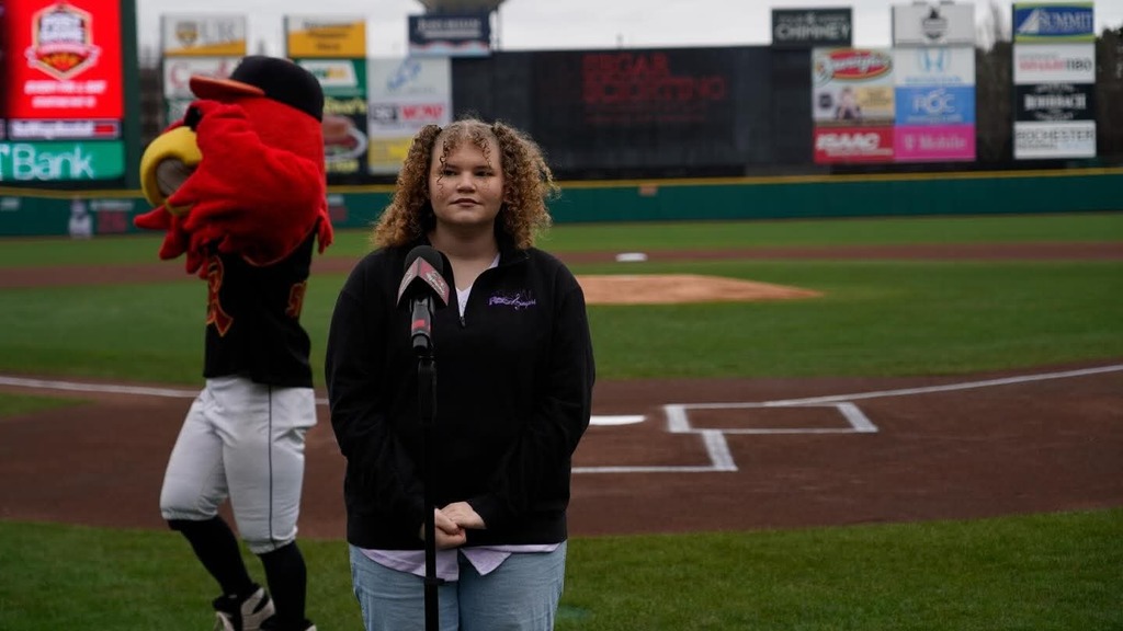 Ella C. stands at mic on baseball field with team mascot in the background