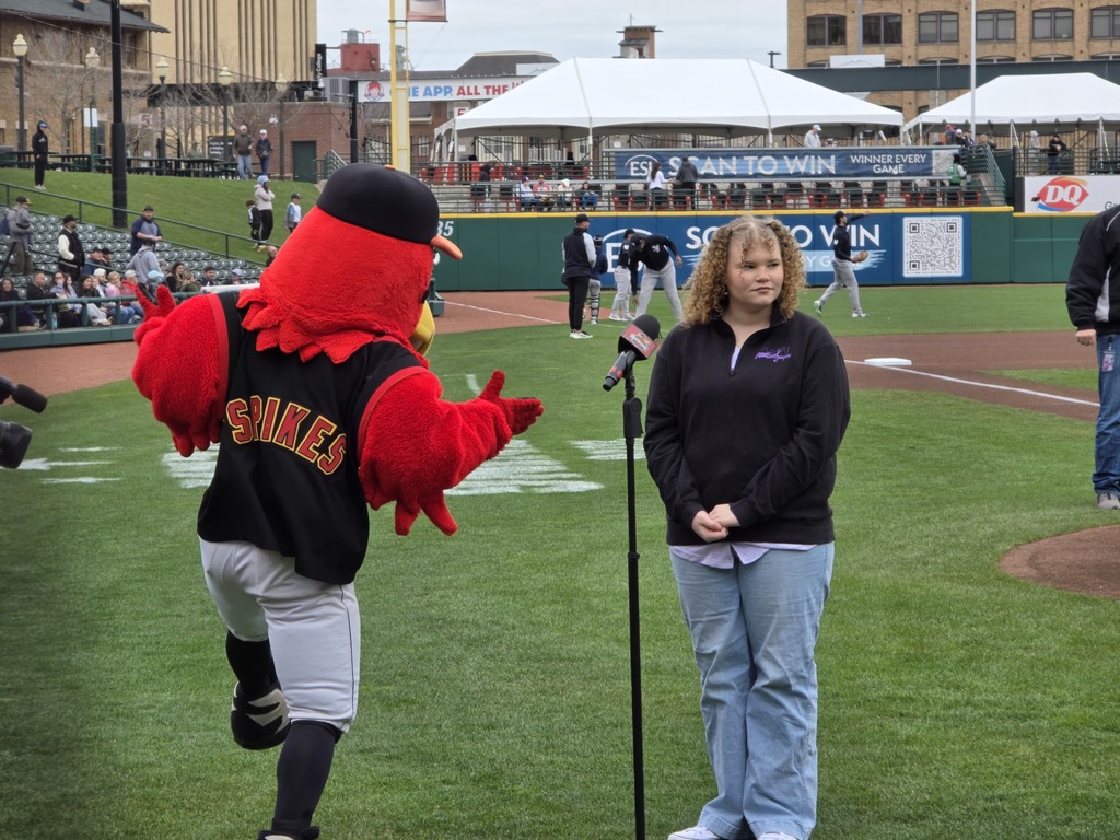 Ella stands at mic stand with team mascot, baseball players, and fans in the back