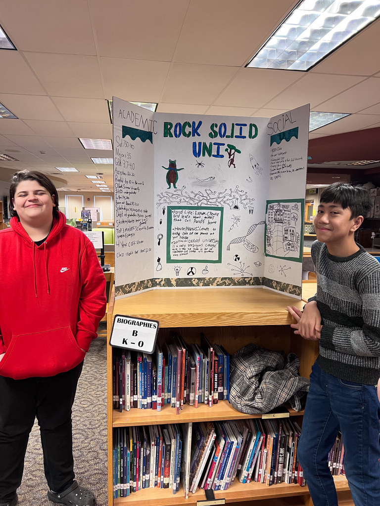 two students stand and smile with presentation board of created university called rock solid uni