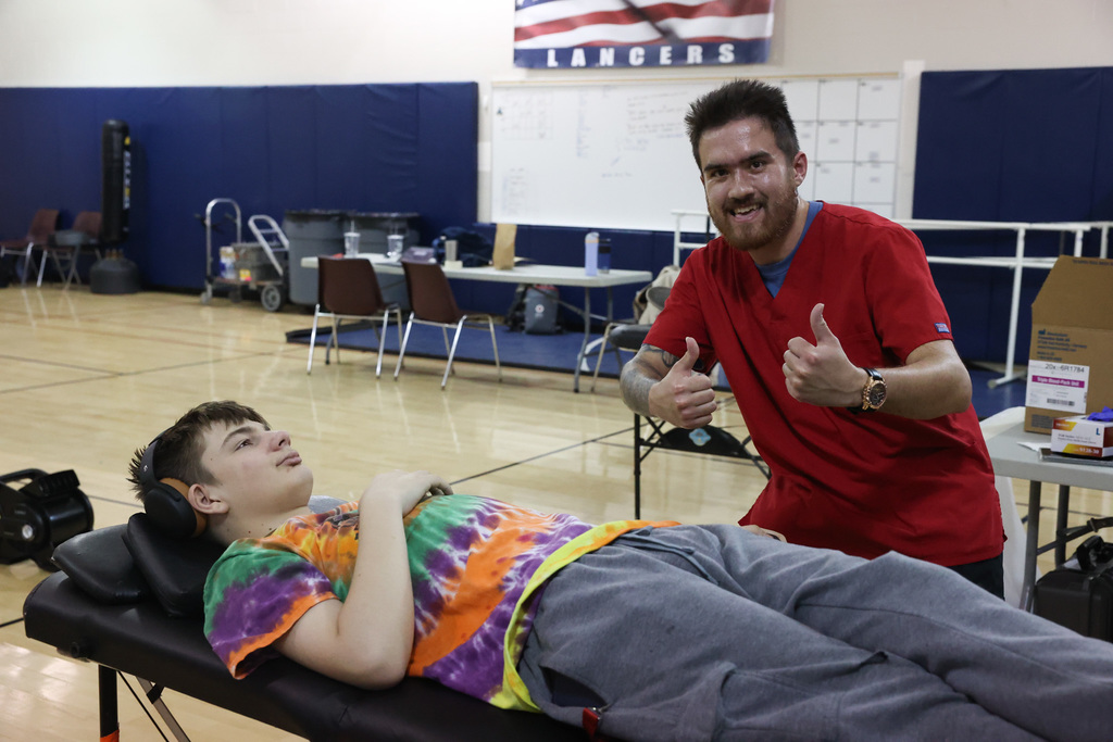 students smiles and sits during blood drive while medical staff puts thumbs up sign