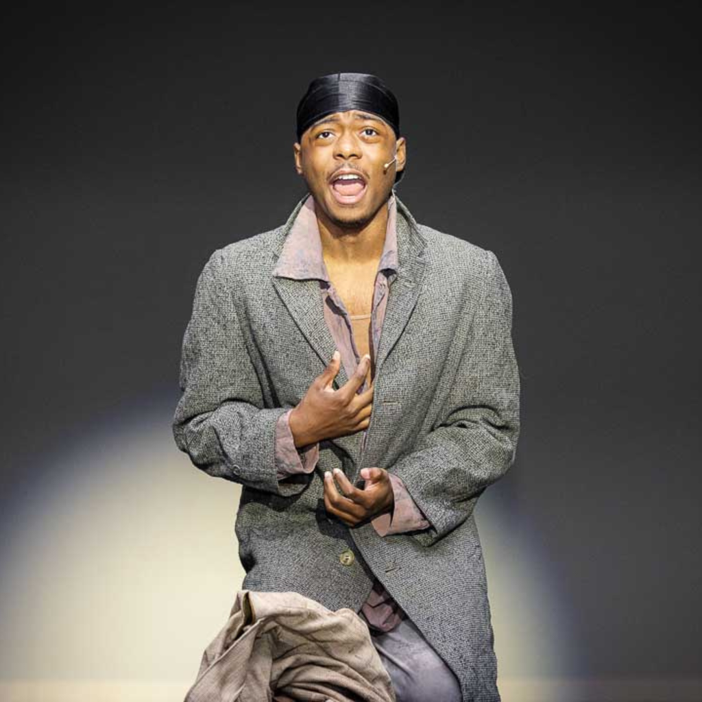 Students sings in a gray coat on stage during a performance of Les Mis