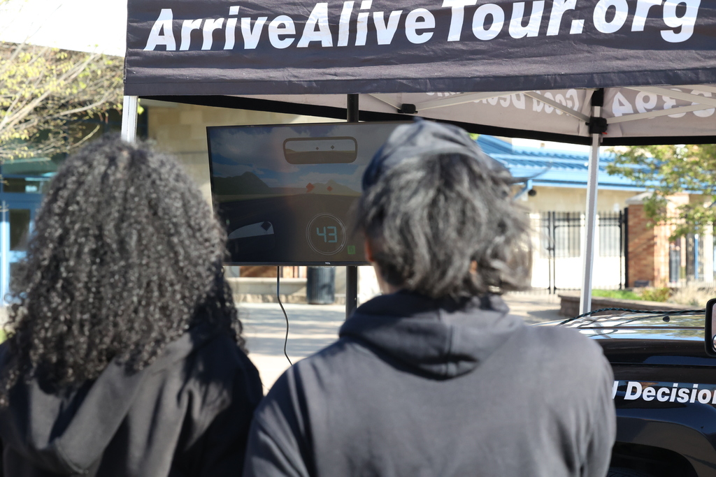 Students watch driving simulation on screen with words on tent ArriveAliveTour.org