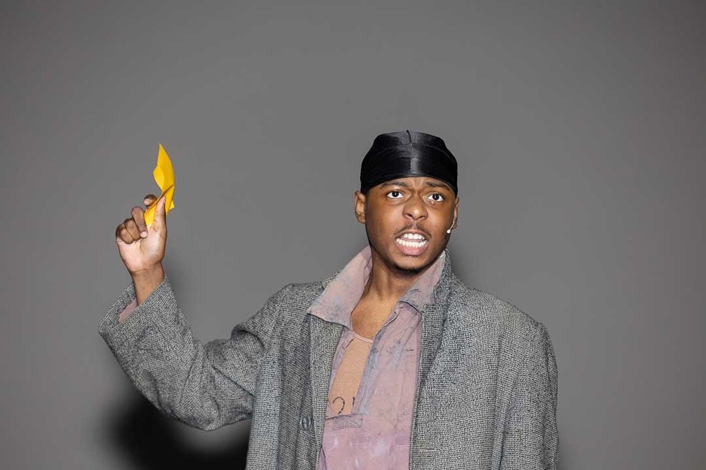 performer holds ticket in his hand and wears gray coat while singing