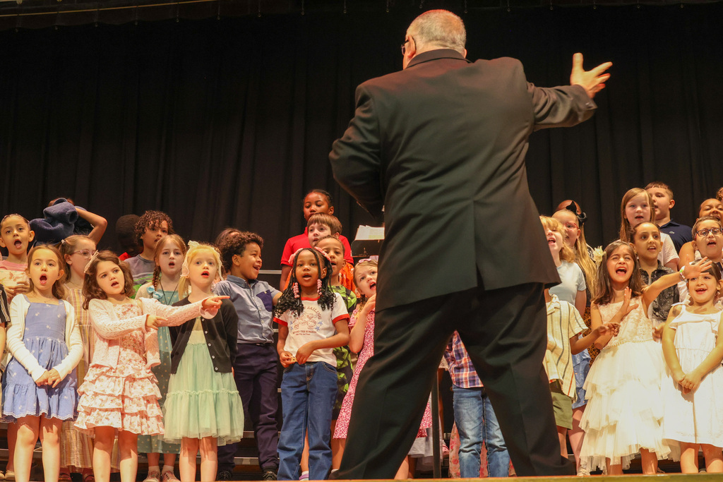 conducter stands and puts arm out to show dance move while students participate on stage