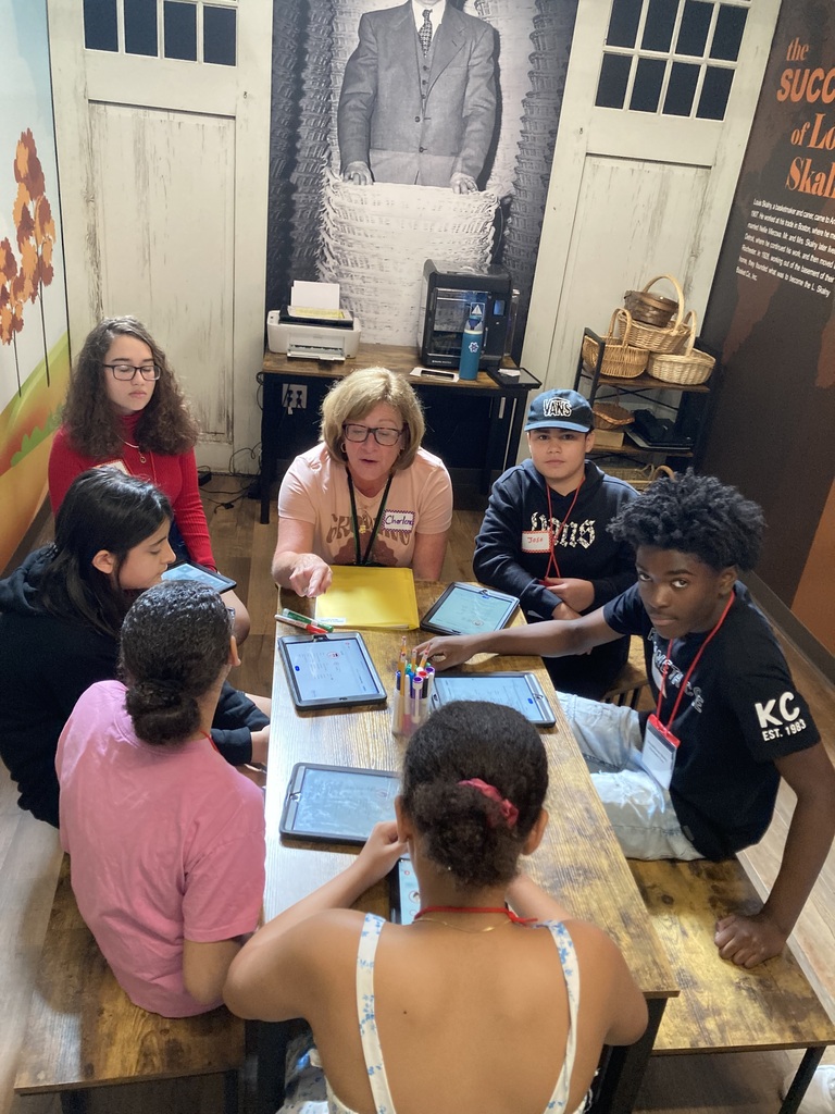 Students work with staff member on field trip at a table with tablets