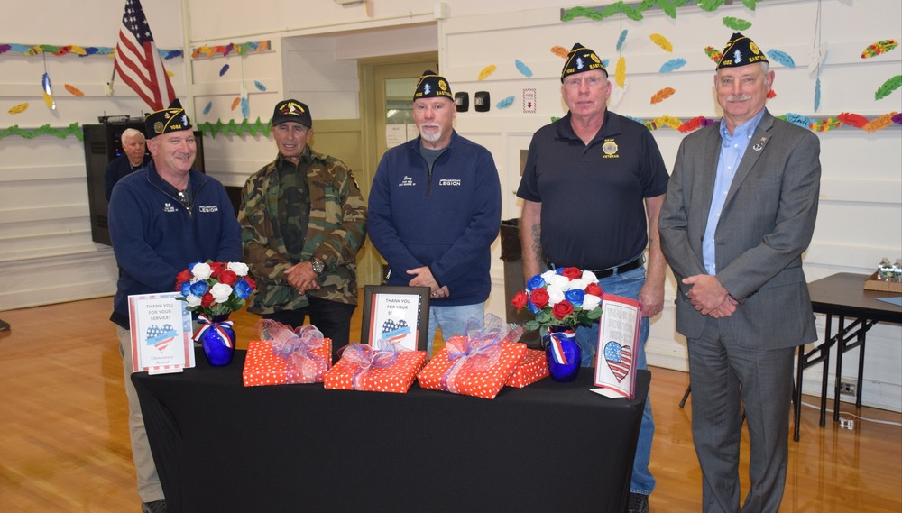 Five men in military hats stand behind a table with gifts and flowers wrapped in patriotic colors. American flag and festive decor are in the background.
