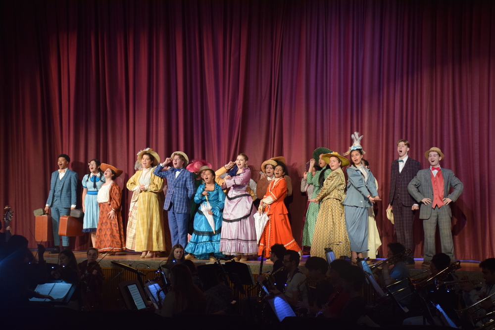 Theatrical performance with actors in colorful, vintage costumes and hats, standing in front of a red curtain. 