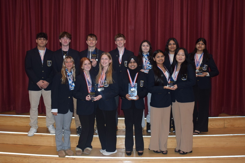 A group of students in matching blazers stand on wooden steps against a red curtain. They hold trophies and wear medals, smiling proudly.