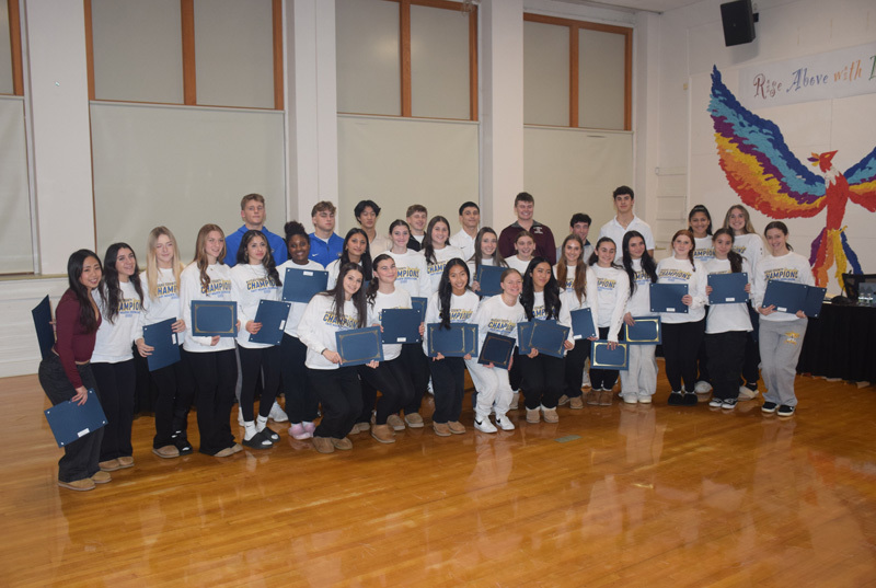 A large group of smiling students poses indoors, holding certificates. They wear matching "champions" shirts. A colorful mural is on the right wall.