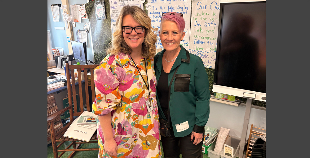 Two women stand smiling in a classroom. One wears a colorful floral dress and glasses, the other a green jacket with pink hair. The setting is warm and welcoming.