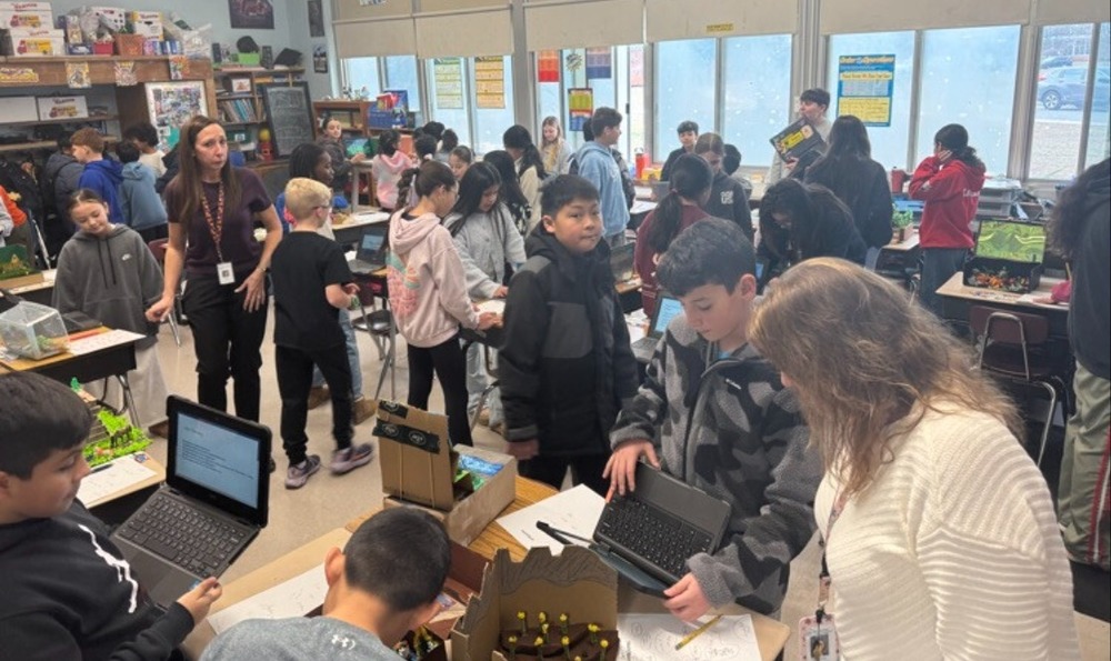 A bustling classroom filled with students and a teacher engaging in various activities. Some students use laptops, while others chat or examine projects, creating an energetic and collaborative atmosphere.