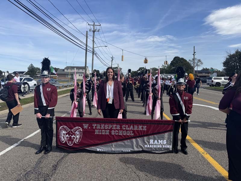 Students marching through street in band attire, holding high school banner.
