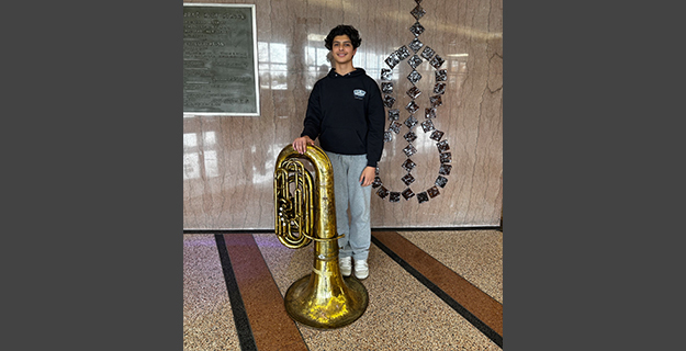 Teen in a black hoodie and gray sweatpants stands indoors, smiling, next to a large brass tuba.