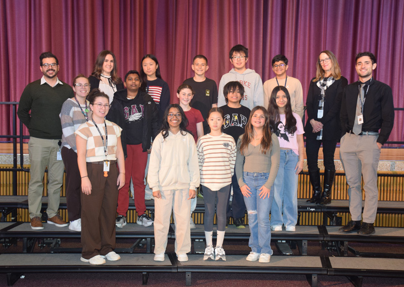 A diverse group of teenagers and adults stand on risers against a purple curtain backdrop, smiling warmly, conveying a sense of camaraderie and community.