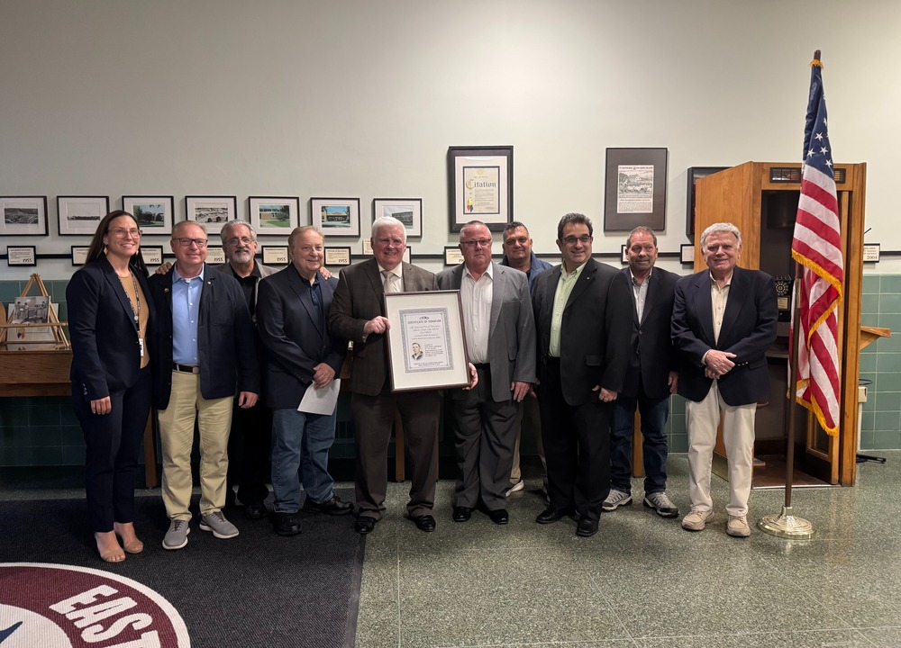 Group photo of community leaders and officials holding a certificate, gathered in a hall with framed photographs and an American flag in the background, celebrating a special recognition event.