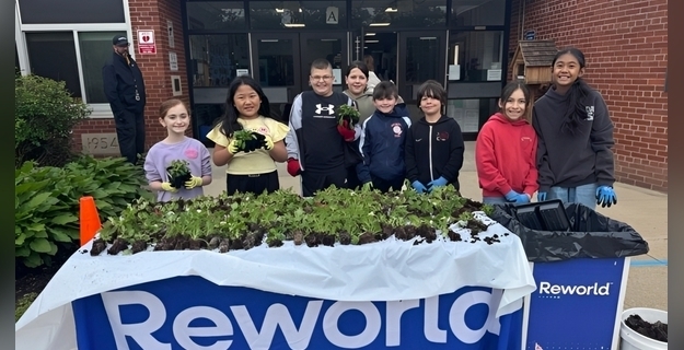 Group of children and adults participating in a community garden project, smiling and holding plants outdoors on a sunny day.