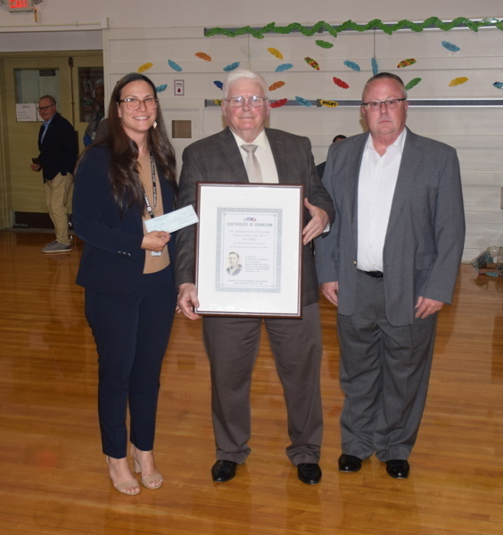 Three individuals at a recognition event, with one woman presenting a check and a framed certificate to a man, while another man stands beside them, all set in a school gymnasium.