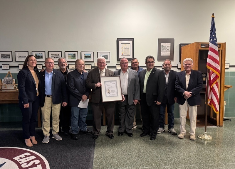 Group photo of community leaders and officials holding a certificate, gathered in a hall with framed photographs and an American flag in the background, celebrating a special recognition event.