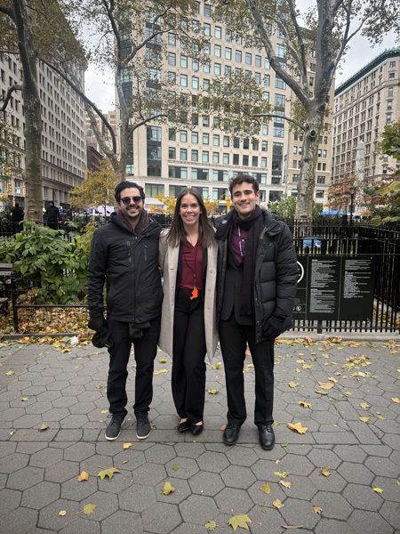 A marching band in red uniforms practices in a park, surrounded by autumn trees and city buildings. Flags are displayed nearby.