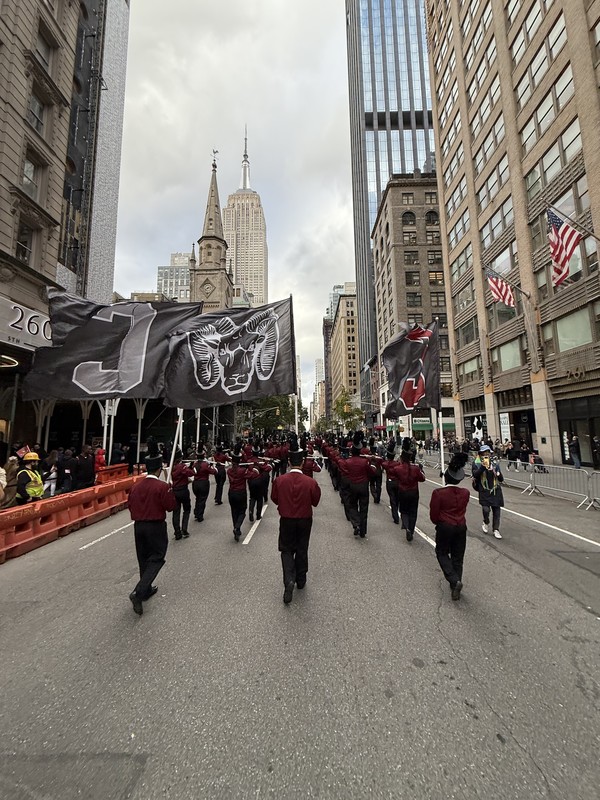 A marching band in red uniforms practices in a park, surrounded by autumn trees and city buildings. Flags are displayed nearby.