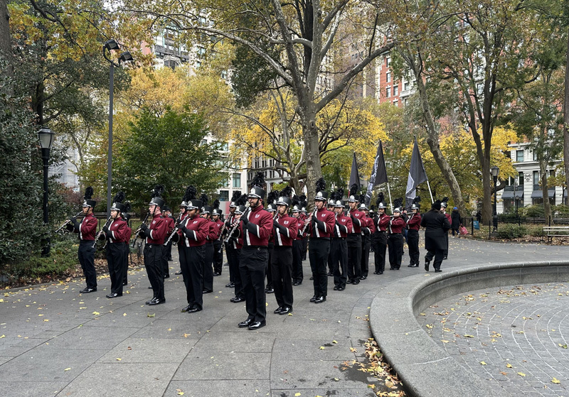 A marching band in red uniforms practices in a park, surrounded by autumn trees and city buildings. Flags are displayed nearby.