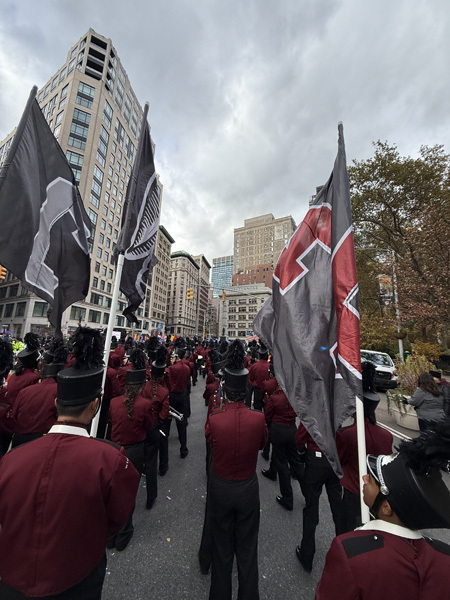 A marching band in red uniforms practices in a park, surrounded by autumn trees and city buildings. Flags are displayed nearby.