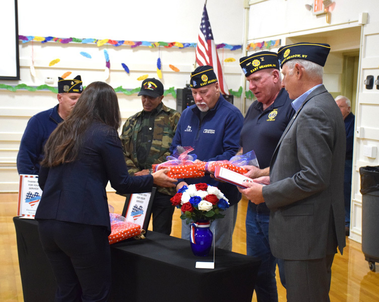 Five men wearing veterans’ caps stand behind a table with patriotic-themed gifts and flowers. 