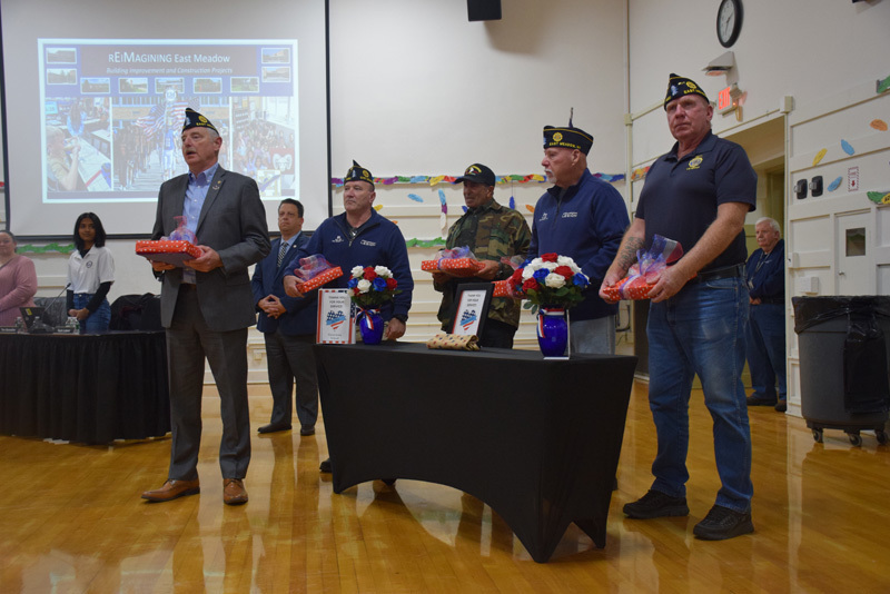 Five men wearing veterans’ caps stand behind a table with patriotic-themed gifts and flowers. 