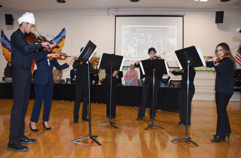 A group of musicians, dressed in formal attire, play violins with sheet music on stands.