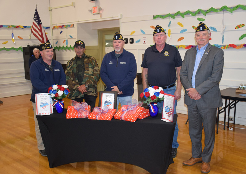 Five men wearing veterans’ caps stand behind a table with patriotic-themed gifts and flowers. An American flag hangs in the background. 