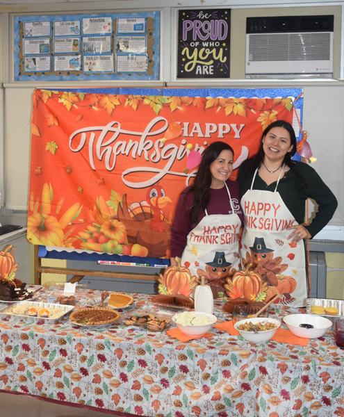Two women smiling and posing in front of a festive "Happy Thanksgiving" banner. They wear themed aprons, surrounded by a table with Thanksgiving foods.